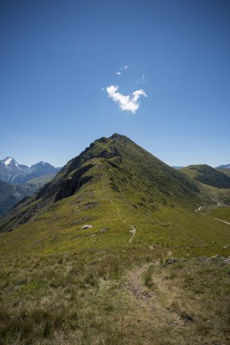 Path going to a green mountain druing summer with a cloud above it and clear blue sky. Alps, Col de Sarenne, Isere, France